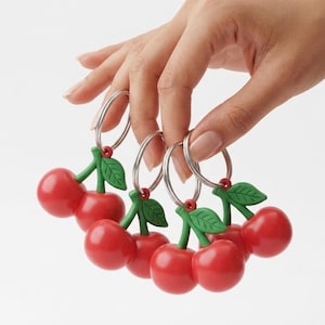 May include: Several red cherry keyrings with green stems and leaves, each attached to a silver metal ring. The keyrings are held up by a hand against a white backdrop.