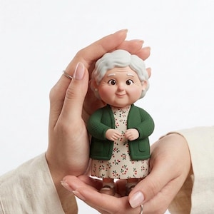 Könnte beinhalten: Figur einer lächelnden älteren Frau mit weißem Haar, die einen grünen Cardigan über einem geblümten Kleid trägt. Die Figur wird in offenen Händen vor weißem Hintergrund gehalten. Die Figur hat rosige Wangen und einen freundlichen Ausdruck.
