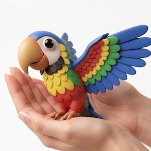 May include: A colorful parrot figurine with outstretched wings, featuring a blue head and wings, a yellow, red, and green body, and a tan beak. The parrot is held in open hands against a white background.