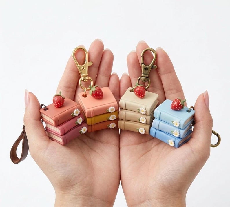 May include: Four miniature book keychains in shades of pink, yellow, beige, and blue, each topped with a red strawberry and small white flowers. The keychains have gold-colored clasps and are held in open hands.