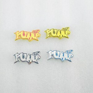 May include: Four metal pins with the word "PUMP" in a graffiti style. The pins are in gold, yellow, silver, and blue. The pins are on a white surface.