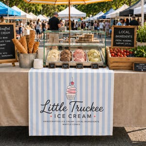 Op de afbeelding: Een ijskraam met een blauw-wit gestreept tafelkleed met de tekst Little Truckee Ice Cream. Een glazen vitrine bevat vier smaken ijs, terwijl een metalen emmer ijshoorntjes bevat en een houten kist verse ingrediënten bevat.
