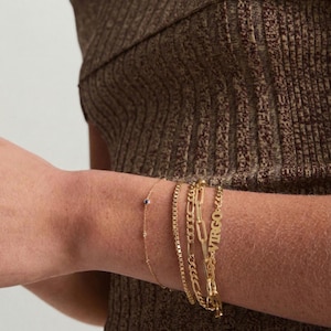 May include: A collection of gold bracelets on a wrist. The bracelets include a delicate chain with a small blue stone, a box chain, a curb chain, and a bracelet with the word "VIRGO".