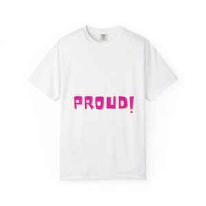 Puede incluir: Camiseta blanca con la palabra "PROUD!" en rosa brillante, delineada en rosa más oscuro. La camiseta tiene cuello redondo y mangas cortas. El texto está centrado en la parte delantera.