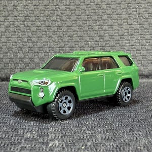 Puede incluir: Un SUV de juguete de metal fundido en color verde lima que presenta un emblema de Toyota en la parrilla delantera, ventanas tintadas oscuras, parachoques de plástico negro y ruedas negras con un diseño de seis radios y detalles moldeados.