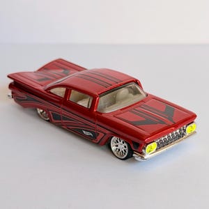 Pode incluir: Um carro de brinquedo vermelho metálico com gráficos de listras pretas no capô, teto e laterais. O veículo possui faróis amarelos brilhantes, uma grade cromada e rodas prateadas de vários raios. Um pequeno logotipo Hot Wheels é visível.