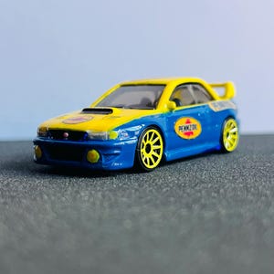May include: A blue and yellow toy car with the Pennzoil logo. The car has a spoiler and yellow wheels. The car is on a gray surface.