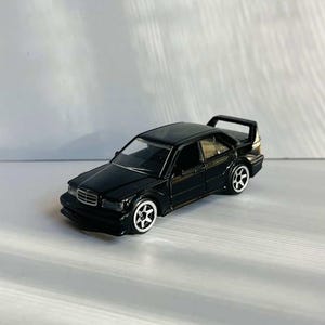 May include: A black die-cast model car with a rear spoiler and white-rimmed wheels. The miniature vehicle has a sleek design and is set against a plain white background. The car's details are clearly visible, showcasing its sporty features.