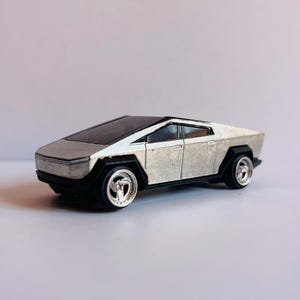 Puede incluir: Un coche de juguete plateado y negro con un diseño futurista. El coche tiene una carrocería plana y angular, neumáticos negros y llantas plateadas. El techo es negro y las ventanas están tintadas. Es una réplica en miniatura de un Cybertruck.