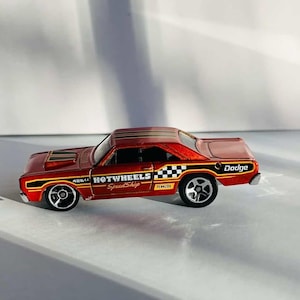 以下が含まれることがあります： 赤と金のHot Wheels Dodgeのおもちゃの車。車には白黒の市松模様の旗と「Hot Wheels Speed Shop」と「Pennzoil」の文字がプリントされています。車は黒いホイールです。