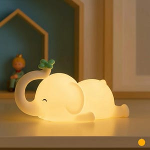 May include: A soft, glowing white elephant-shaped night light with a curved trunk and a small green leaf on top. The elephant is lying down, with a smiling face. The light is on, illuminating the room.