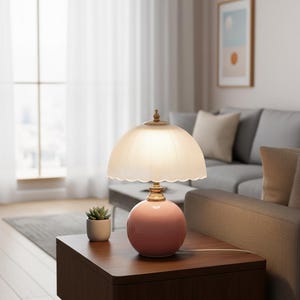 May include: A pink ceramic table lamp with a scalloped, frosted glass shade. The lamp has a brass base and a decorative finial. A small potted succulent sits nearby on a dark wood table. The lamp is a decorative home decor item.