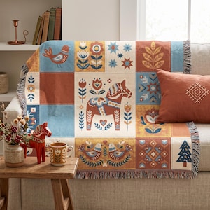 May include: A woven throw blanket featuring a grid of rectangular panels with folk-inspired motifs including a Dala horse, birds, flowers, stars, and a pine tree in shades of terracotta, mustard yellow, cream, and muted blue with fringed edges.