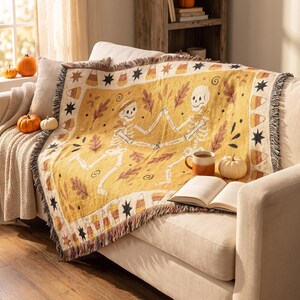Op de afbeelding: Een geweven plaid met een mosterdgeel middenstuk met twee dansende skeletten, bruine herfstbladeren en kleine krullen. De rand toont een herhalend patroon van zwarte sterren en oranje, gele en witte snoepmaïs-vormen.