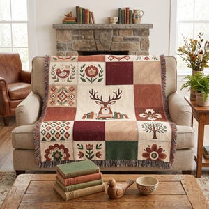 May include: A woven throw blanket featuring a patchwork design with a central deer illustration surrounded by squares containing floral motifs, geometric patterns, a bird, and a tree in shades of cream, burgundy, rust, green, and brown.