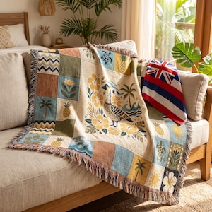 May include: A woven throw blanket featuring a patchwork design with tropical motifs including a nene goose, hibiscus flowers, palm trees, pineapples, sea turtles, and waves, accented by a folded flag with red, white, and blue horizontal stripes.