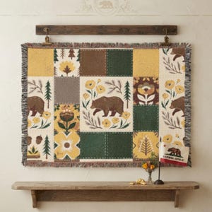 Könnte beinhalten: Eine gewebte Wandteppichdecke mit einem Patchwork-Design aus Bären, Tannen und gelben Blumen in Grün-, Braun- und Goldtönen. Die Decke hat einen Fransenrand und hängt an einem dunklen Holzbügel. Eine kleine Flagge von Kalifornien ist zu sehen.