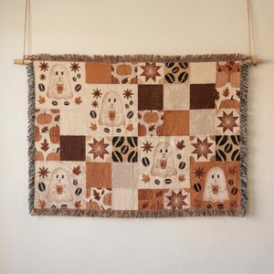Puede incluir: Un tapiz tejido rectangular colgado de una varilla de madera, con un diseño de patchwork en tonos marrón, crema y beige. El patrón incluye ilustraciones de fantasmas sosteniendo tazas de café, calabazas, granos de café y hojas en forma de estrella.