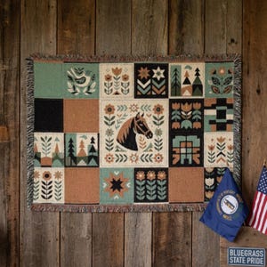 Può includere: Una coperta tessuta con un design patchwork caratterizzato da un motivo centrale a testa di cavallo circondato da motivi floreali, alberi e geometrici nei toni del verde, marrone, nero e crema, rifinita con un bordo sfrangiato su tutti i lati.