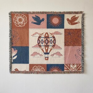 May include: A square woven tapestry blanket featuring a central hot air balloon surrounded by a grid of panels. The design includes illustrations of birds, suns, and flowers in a color palette of terracotta, navy blue, pink, and cream with fringe.
