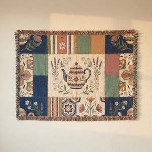 May include: A rectangular woven tapestry featuring a central cream-colored panel with a teapot illustration surrounded by lavender and daisies. The border consists of patchwork-style blocks in navy blue, sage green, and muted orange with floral motifs.