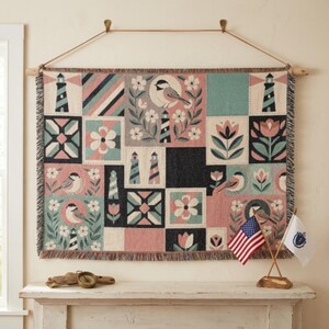 May include: A woven wall tapestry featuring a patchwork design of lighthouses, chickadees, and floral motifs in muted shades of pink, teal, and black. The rectangular textile has a fringed bottom edge and is suspended from a wooden rod with a cord.