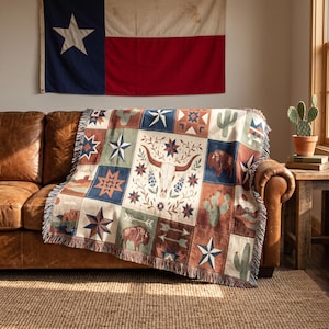 May include: A woven throw blanket featuring a patchwork design with illustrations of a longhorn skull, bison, cacti, and stars in shades of blue, red, and tan. The blanket has a fringed edge and is draped over a brown leather sofa.
