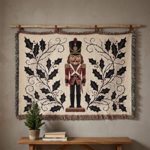 May include: A rectangular woven wall tapestry featuring a central nutcracker figure in a red and black uniform, flanked by dark green holly leaf and berry branches on a cream background, finished with fringed edges and hung from a wooden rod.