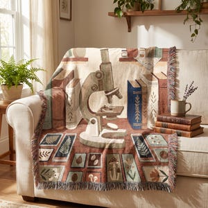 Op de afbeelding: Een geweven plaid met een centrale illustratie van een microscoop, omringd door stapels boeken, botanische specimens en kleine ingelijste natuurmotieven zoals varens, paddenstoelen en bloemen in gedempte aardetinten met een franje.