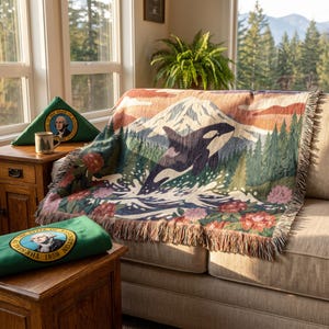 May include: A woven throw blanket featuring a black and white orca jumping from water, a snow-capped mountain, pine trees, and flowers. Two green folded fabrics display the circular Seal of the State of Washington with George Washington's portrait.
