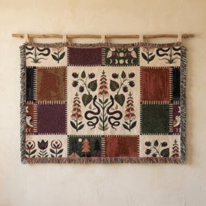 Puede incluir: Un tapiz de pared tejido rectangular con diseño de patchwork y bordes con flecos. El panel central muestra dos serpientes entrelazadas con flores de dedalera. Los paneles circundantes incluyen motivos de árboles, lunas, búhos y patrones florales en tonos tierra.