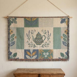 Peut inclure: Une tenture murale tissée rectangulaire présentant une illustration centrale de théière entourée de brins floraux. La tapisserie affiche une grille de panneaux dans des tons vert, crème et jaune, finie avec un bord frangé et une tige en bois.