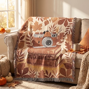 May include: A woven throw blanket featuring a repeating pattern of brown and orange autumn leaves. The center design displays a vintage-style camera resting on a stack of three leather-bound books, all rendered in warm shades of brown and tan.