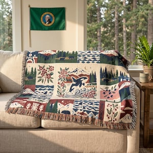 May include: A woven throw blanket featuring a patchwork design of Pacific Northwest motifs including an orca, mountains, pine forests, bears, salmon, and floral elements in shades of navy blue, forest green, cream, and deep red with fringed edges.