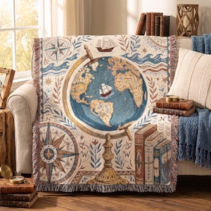 Peut inclure: Un plaid tissé présentant une illustration centrale de globe avec un navire, entouré de motifs nautiques incluant une rose des vents, des vagues stylisées et des livres empilés. Le design utilise une palette de tons bleu, crème et brun avec franges.