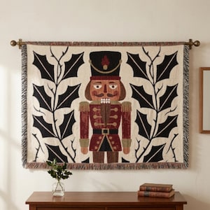May include: A rectangular woven wall tapestry featuring a central nutcracker figure in a red and brown uniform with a black hat. The design is flanked by large black holly leaf patterns on a cream background, finished with a fringed edge.