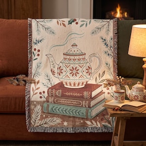 May include: A woven throw blanket featuring a central illustration of a patterned teapot resting on a stack of three books. The design includes surrounding pine branches, berries, and leaves in muted tones of green, red, and cream with fringed edges.