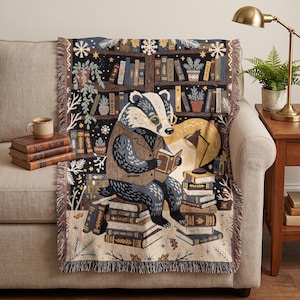 Pode incluir: Uma manta tecida com um texugo usando óculos e colete, sentado numa pilha de livros enquanto lê. O fundo retrata uma estante escura cheia de livros, plantas em vasos e flocos de neve brancos, com acabamento em franjas nas bordas.