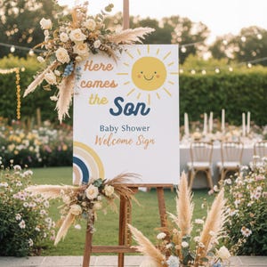 Op de afbeelding: Een houten ezel toont een welkomstbord voor een babyshower met de tekst "Here comes the Son". Het bord heeft een lachende zon en een regenboog. Versierd met witte rozen en pampasgras.
