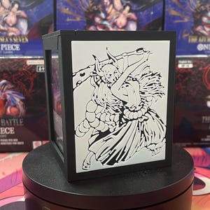 Op de afbeelding: Een zwarte deckbox met een witte reliëf-illustratie van een personage dat een grote knots vasthoudt, geplaatst op een zwarte ronde standaard. De doos staat voor verschillende One Piece kaartspel boosterboxen met zichtbare tekst en afbeeldingen.