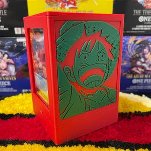 May include: A bright red rectangular card storage box featuring a raised green relief of a cartoon character with a wide-eyed expression and a straw hat. The front panel has a clear window displaying a trading card inside the plastic container.