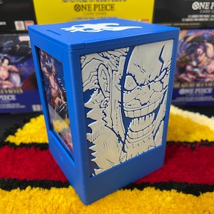 Può includere: Una scatola rettangolare blu per carte con un grafico bianco in rilievo di un personaggio sorridente sul pannello laterale. Il coperchio mostra un emblema bianco con teschio e ossa. Una carta di One Piece è visibile dalla finestra laterale.