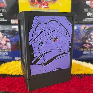 Op de afbeelding: Een zwarte rechthoekige deckbox met een paarse reliëfafbeelding van een anime-personage met een bril en gezichtsbedekking. Het personage heeft één rood oog en één blauw oog. De doos heeft een strak, gestructureerd ontwerp met een matte afwerking.