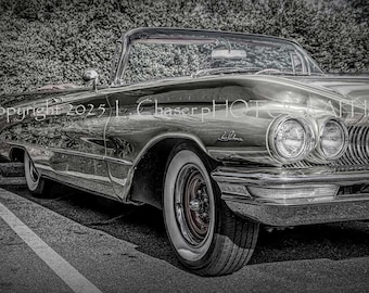 Op de afbeelding: Een klassieke cabriolet met een glanzende chromen afwerking, whitewall-banden en een rode wielnaaf. De auto staat geparkeerd op een asfalt oppervlak met een groene haag op de achtergrond. De afbeelding is in zwart-wit.
