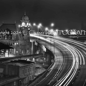 May include: A black and white photograph of a building with a clock tower at night. The building is lit up and there are cars driving on a highway in the foreground. The text "Copyright 2024 L. Chaser Photographs" is visible in the image.