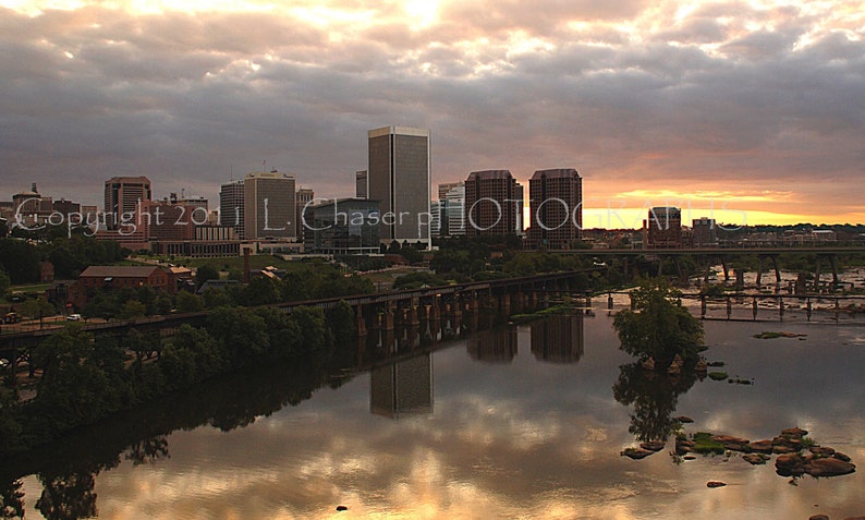 May include: A panoramic view of a city skyline at sunset, with tall buildings reflected in the water. The sky is a mix of orange, pink, and gray clouds. A bridge crosses the river in the foreground.