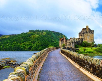 May include: A stone bridge leads to a large, gray stone castle on a small island. The castle is surrounded by green trees and water. The sky is blue with white clouds.