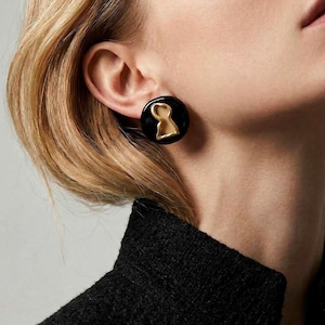 May include: Black and gold keyhole stud earrings. The earrings feature a black circular base with a gold keyhole design. The earrings are worn on the ear of a person wearing a black jacket.