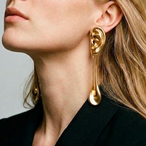Puede incluir: Pendientes dorados con forma de oreja, con una larga extensión en forma de lágrima. Los pendientes los lleva una persona con cabello rubio y una chaqueta negra. Un accesorio único y artístico.