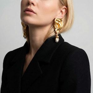 May include: Gold-toned ear-shaped earrings with dangling pearl accents. The earrings are detailed with small embellishments and are worn against a black jacket. The design is artistic and unique, showcasing a statement jewelry piece.